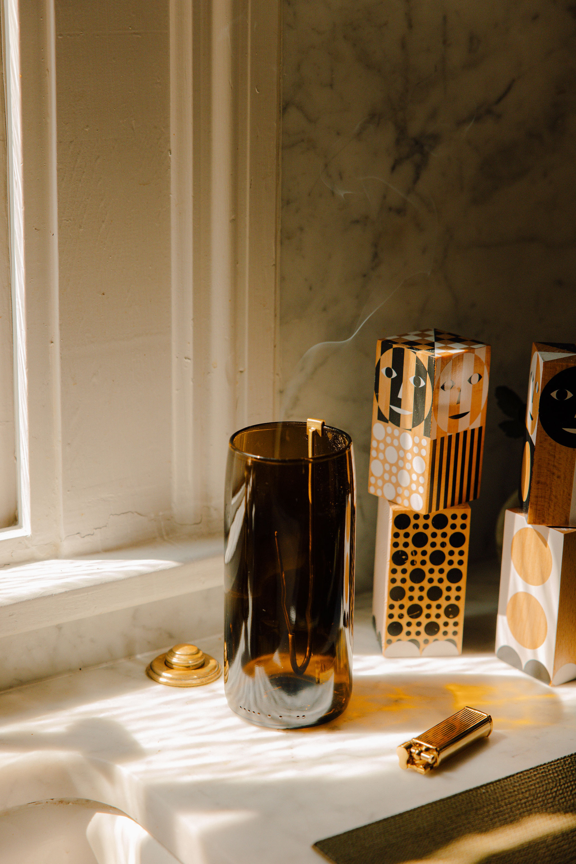 Recycled bottle incense vase with brass hook, incense smoke, and a brass lighter in warm window light.
